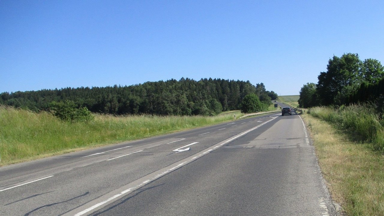 Man blickt auf eine Straße mit beschädigtem Fahrbahnbelag mit Fahrbahnmarkierungen; links und rechts Grünstreifen im Hintergrund ein Wald