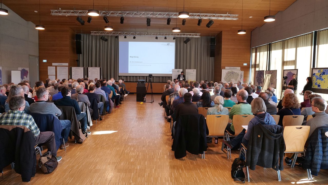 Man blickt von hinten in einen voll besetzten Veranstaltungsraum. Ganz vorne ist das Plenum mit einem Sprecher zu erkennen.