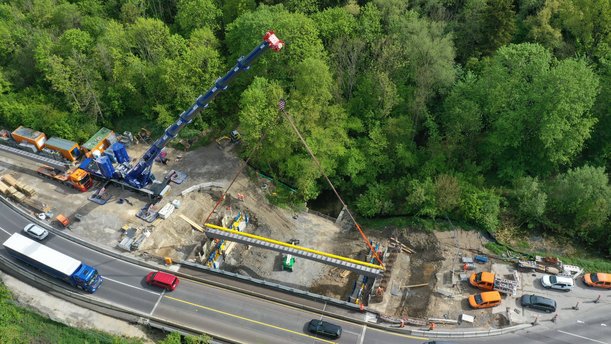 Luftbild einer Baustelle. Ein langer STahlträger wird von einem Kran angehoben, daneben läuft eine Straße