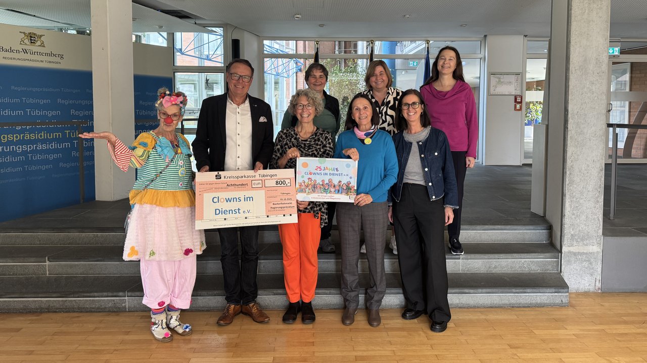 Das Foto zeigt wie Regierungspräsident Klaus Tappeser gemeinsam mit dem Organisationsteam sowie den helfenden Händen des Bücherflohmarkts den Scheck an Christel Ruckgaber übergibt. Ein Clown ist ebenfalls auf dem Foto zu sehen.