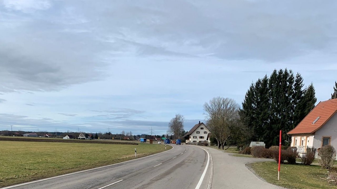 Das Foto zeigt eine Straße. Links eine große Wiesenfläche und rechts und weiter im Hintergrund mehrere Gebäude