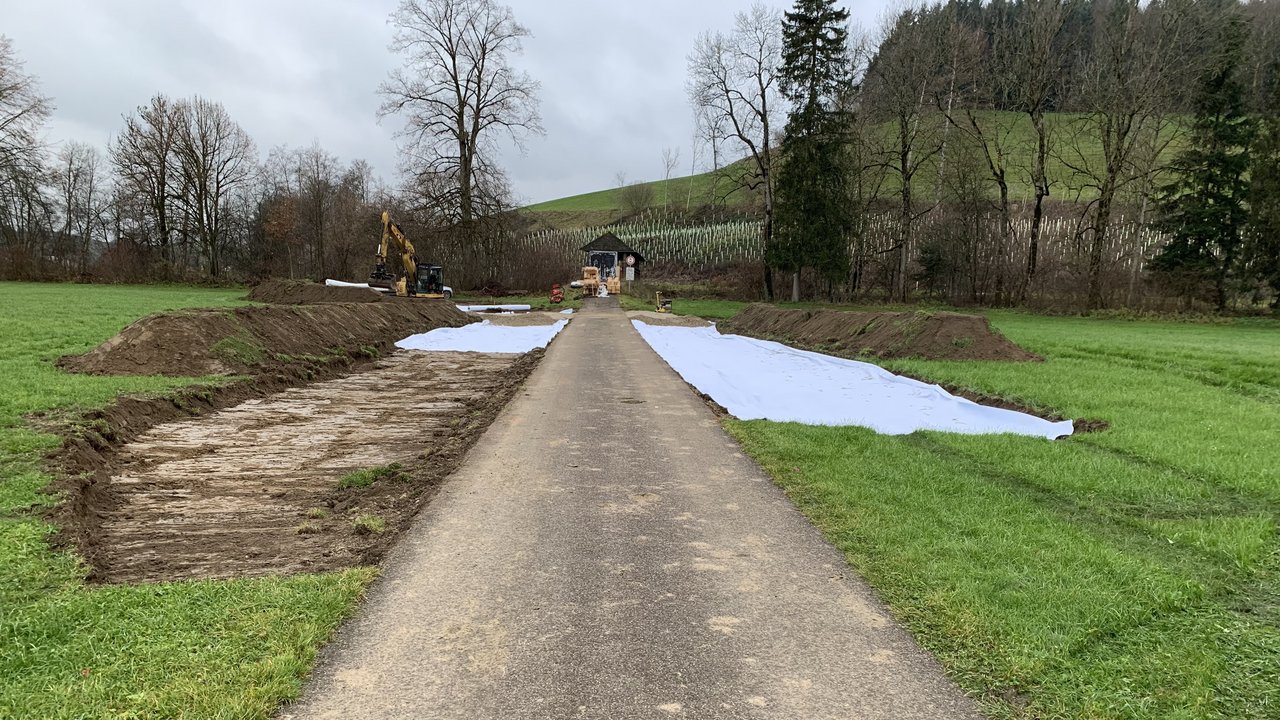 Das Bild zeigt eine Straße, die auf eine Holz-Radwegbrücke zuläuft; links und rechts Wiesen und im Hintergrund ein Bagger und Bäume