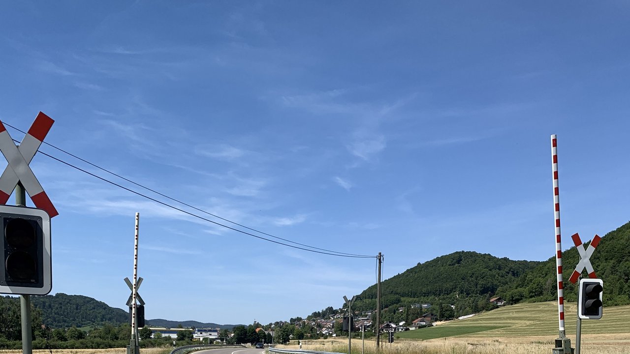 Man blickt auf die Fahrbahn der B 32 vor einem Bahnübergang; Schranken; Andreaskreuz; Ampel; im Hintergrund eine Ortschaft und Wälder; links 