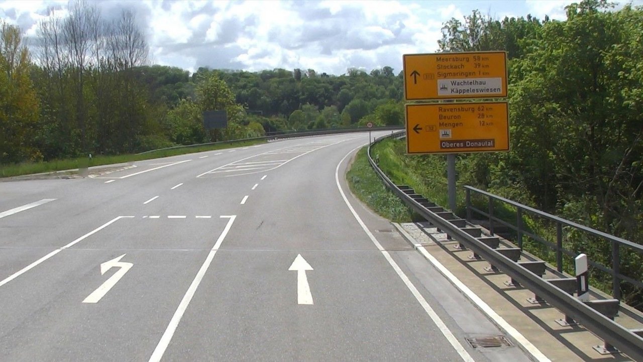 Das Foto zeigt eine 2-spurige Straße mit Straßenmarkierungen und Abbiegespur; Straßenschild und Leitplanken rechts