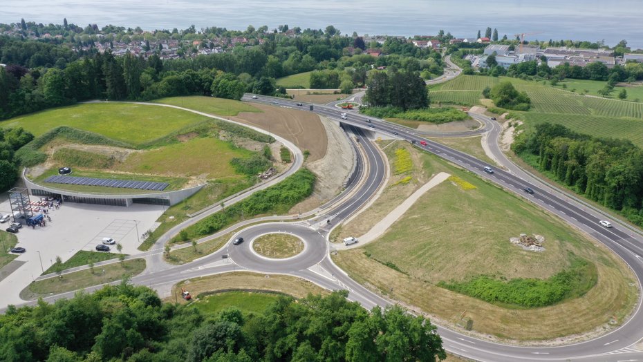 20. Mai 2025: Die fertige Maßnahme kurz vor der Öffnung für den Verkehr. Luftbild einer Straße. Im Hintergrund ist der Bodensee und eine Siedlung zu erkennen. Im Vordergrund ein Kreisverkehr, von dem eine Zufahrt auf die große Straße führt.