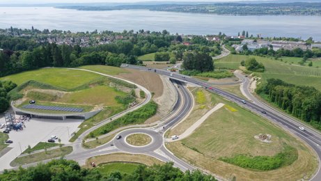 20. Mai 2025: Die fertige Maßnahme kurz vor der Öffnung für den Verkehr. Luftbild einer Straße. Im Hintergrund ist der Bodensee und eine Siedlung zu erkennen. Im Vordergrund ein Kreisverkehr, von dem eine Zufahrt auf die große Straße führt.