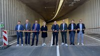 8 Personen stehen nebeneinander in einem Tunnelportal und blicken in Richtung der Kamera. Vor ihnen ist ein Absperrband in den Farben Schwart, Rot und Gold gespannt, an dem sie gerade mit Scheren ansetzen.