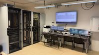 Ein Raum mit mehreren Serverschränken. An einer Wand hängt ein großer Monitor über einem Schreibtisch, auf dem mehrere kleine Monitore stehen.