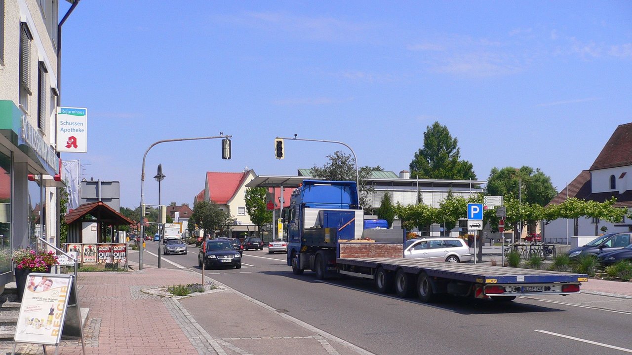 Man sieht die Ortsdurchfahrt von Meckenbeuren, eine Ampelanlage, die mit mehreren Fahrzeugen befahrene B 30, links eine Apotheke und andere Geschäfte und rechts mehrere Gebäude