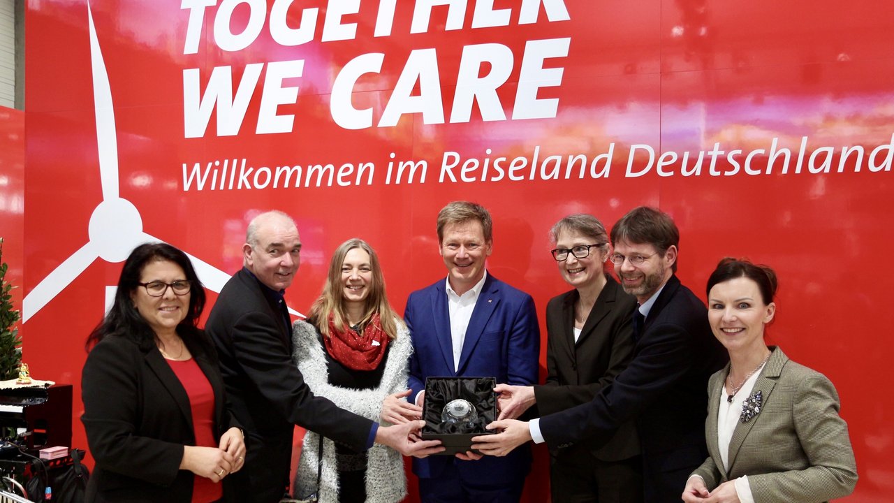 Vier Frauen und drei Männer stehen vor einem roten Plakat; alle zusammen halten den Ehrenpreis - eine Bleikristallkugel - in den Händen