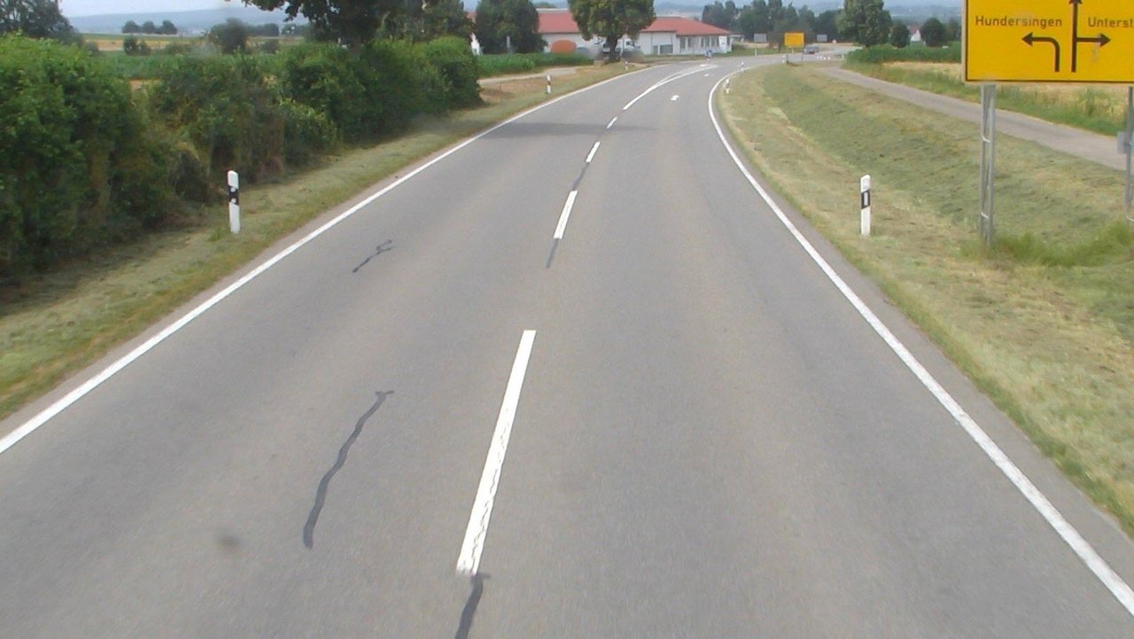Das Foto zeigt eine einspurige Straße mit Fahrbahnmarkierung. Leitplanken. Links und rechts Grünstreifen und ein gelbes Straßenschild