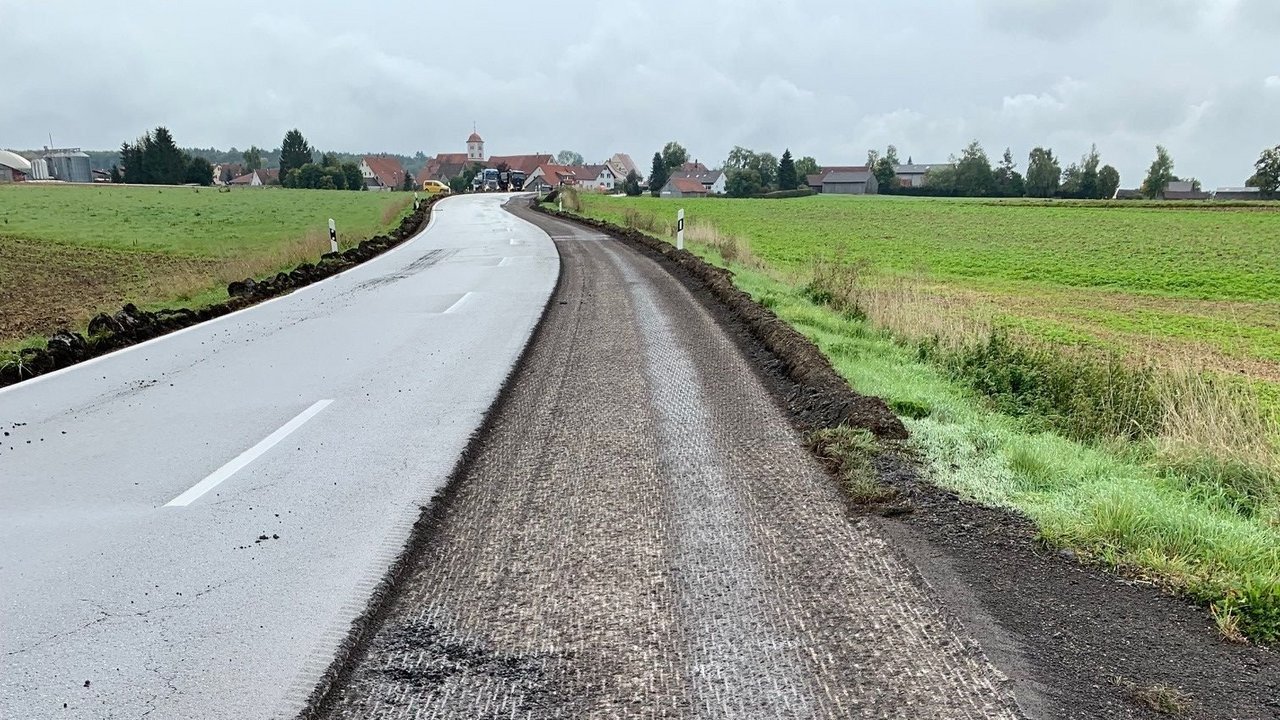 Das Bild zeigt die ausgerissene Fahrbahn der B 312 die erneuert werden soll; links und rechts Wiesen; im Hintergrund ein Dorf