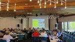 Eine Gruppe Menschen sitzt in einem Saal, an den vordersten Tischen sitzen mit Blick zum Publikum 4 Menschen. Hinter diesen 4 ist eine Leinwand mit einer Präsentation zu sehen.