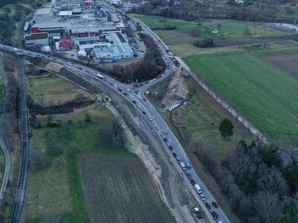 Luftbild einer Straßenbaustelle. Neben dem bestehenden Straßendamm ist Erde aufgeschüttet, wodurch der Straßendamm breiter wird. 
