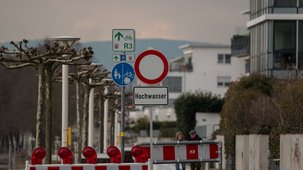 Ein Straßenschild verweist auf Hochwasser, die Straße ist abgesperrt, im Hintergrund sieht man WohnbebauungHintergrund ist 