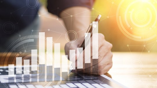 Ein Mann arbeitet am PC vor gelbem Hintergrund, man sieht Wirtschaftssymbole