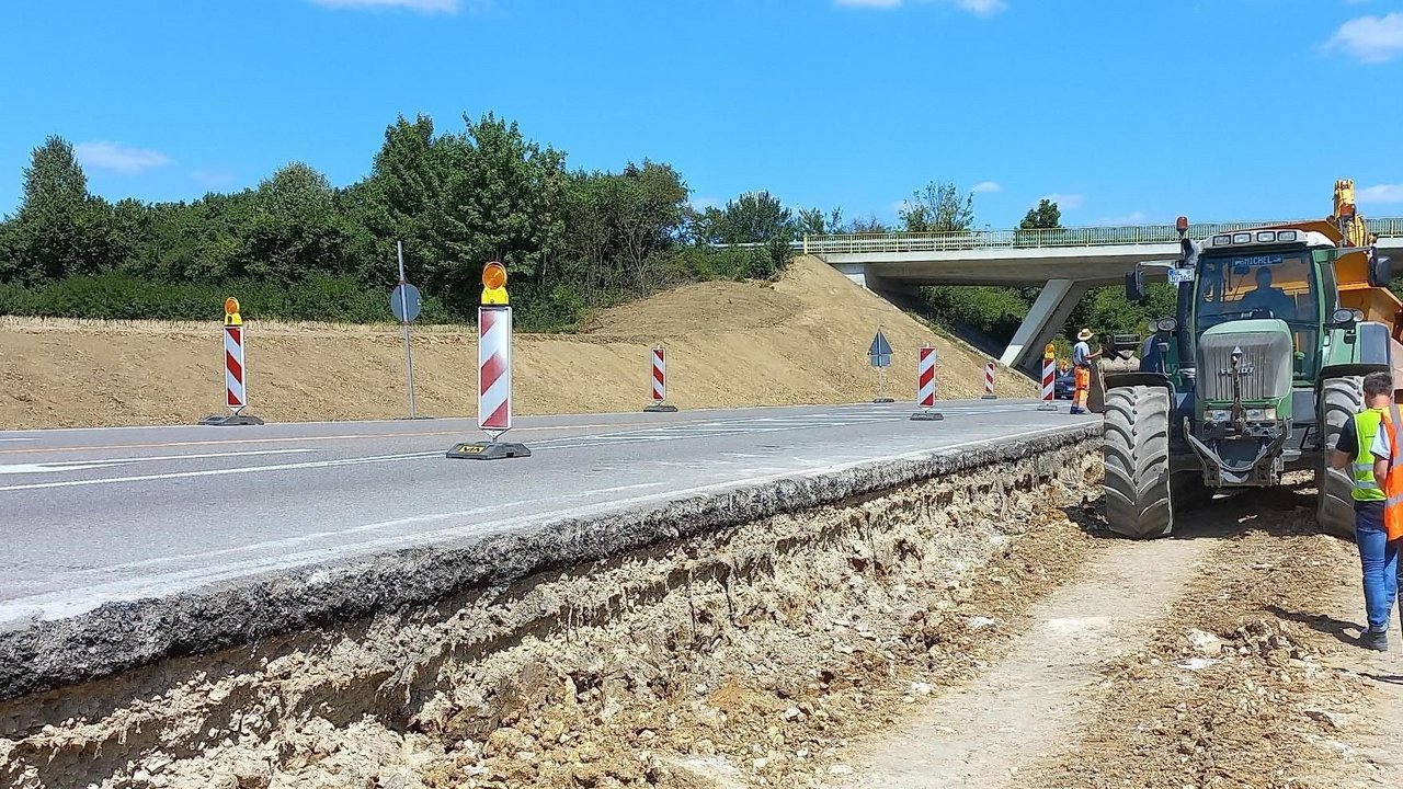 Man sieht die Baustelle/Belagsarbeiten zur Verbreiterung der L 1230 für Ein- und Ausfädelstreifen, Blickrichtung Nord, ein Baustellenfahrzeug und im Hintergrund eine Brücke