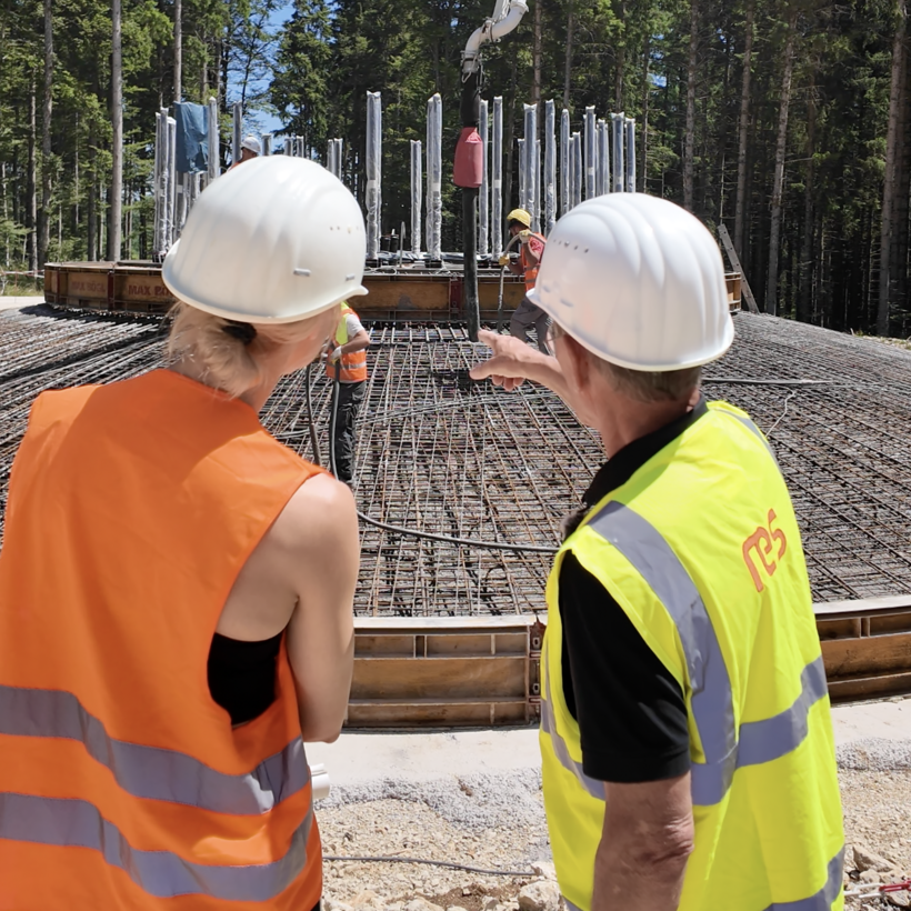 Beobachtung Beton in Fundament Rückenansicht einer Frau und eines Mannes, die auf die Baustelle und die Anlieferung des Betons schauen. Der Mann zeigt auf ein interessantes Detail.