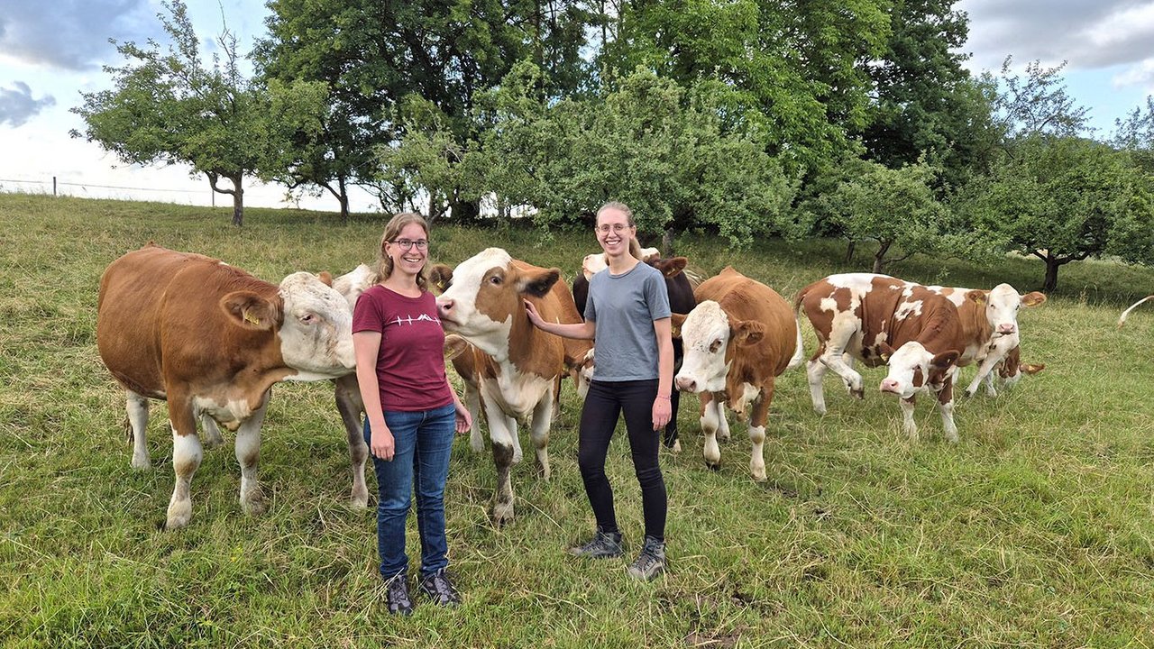 Bild Team Tierwohl Dr. Miriam Knauer und Anna Sulz auf einer Kuhweide mit braunen Kühen mit Bäumen im Hintergrund