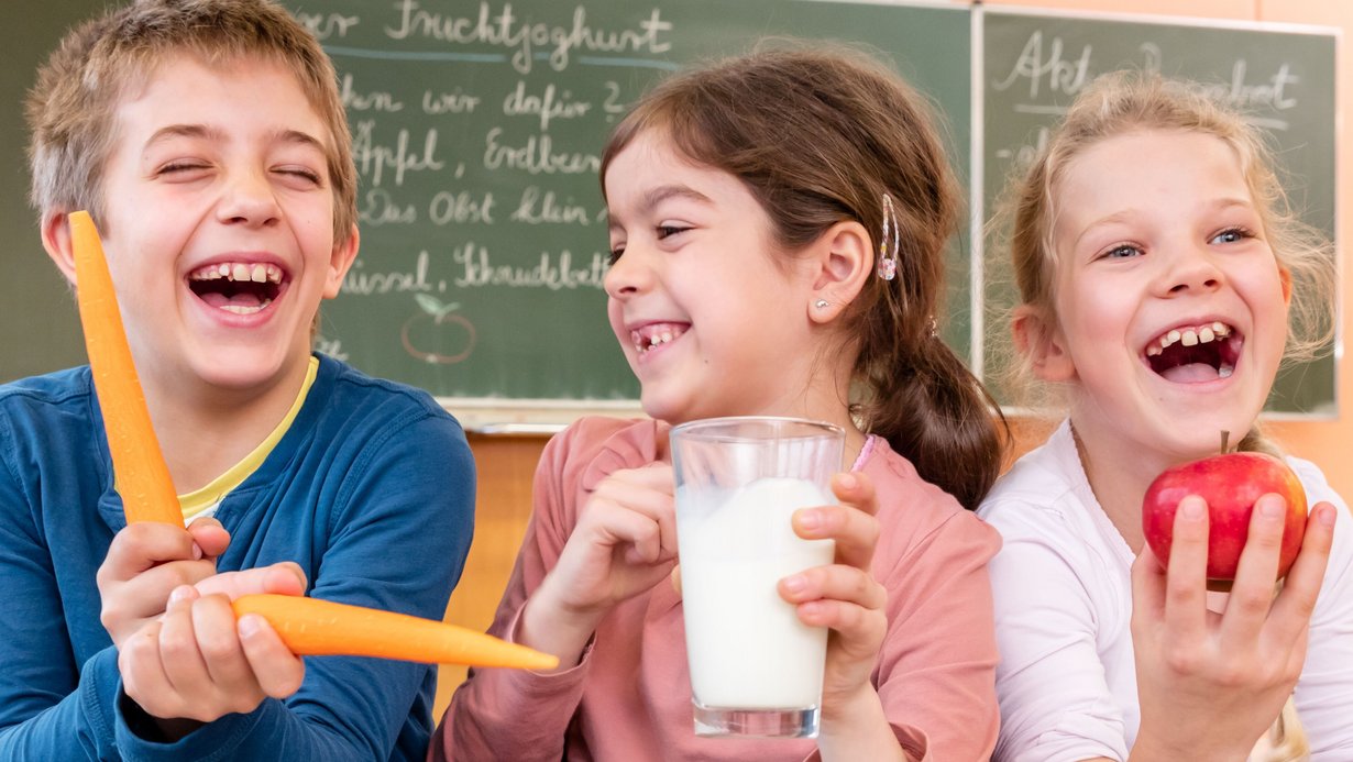 Eine Junge und zwei Mädchen sitzen nebeneinander und lachen in die Kamera, der junge spielt mit zwei Karotten, das Mädchen in der Mitte hält einen Apfel und das Mädchen daneben ein Glas Milch in der Hand