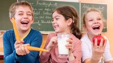 Eine Junge und zwei Mädchen sitzen nebeneinander und lachen in die Kamera, der junge spielt mit zwei Karotten, das Mädchen in der Mitte hält einen Apfel und das Mädchen daneben ein Glas Milch in der Hand