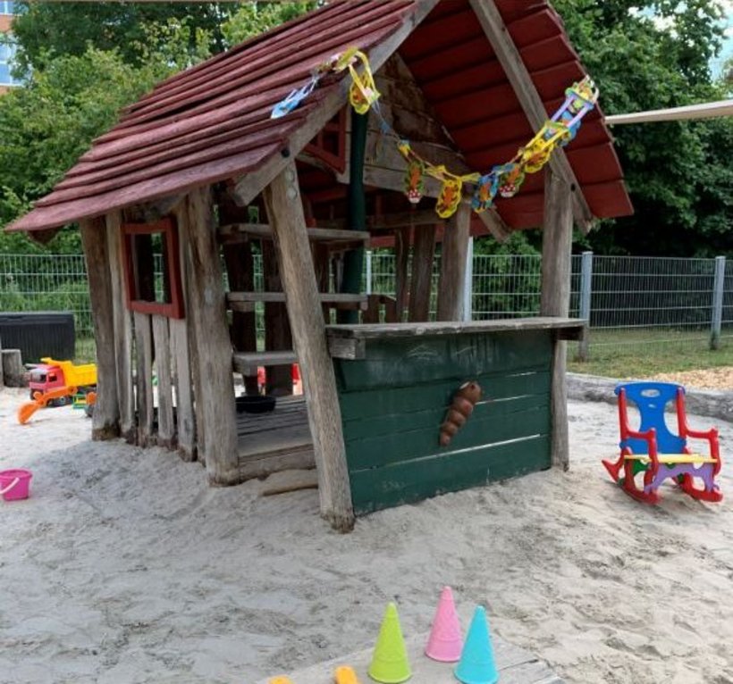 Blick auf eine Spielhütte für Kinder aus Holz, davor sind Sandelspielzeuge