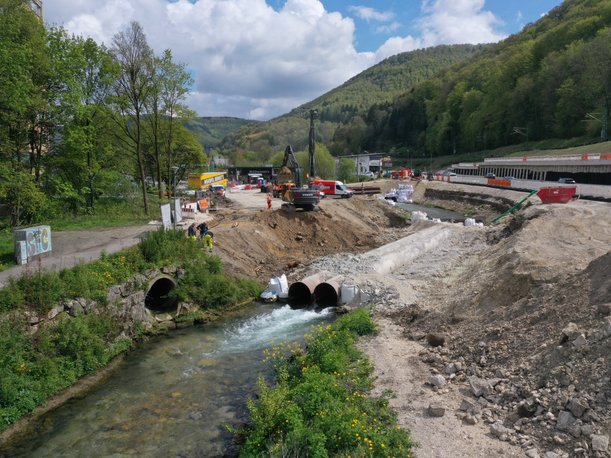 Foto eines Flusses, in dem zwei Stahlrohre liegen, durch die das Wasser fließt. An beiden Böschungen enden Straßenabschnitte, Bagger und Baumaschinen stehen in der Nähe.